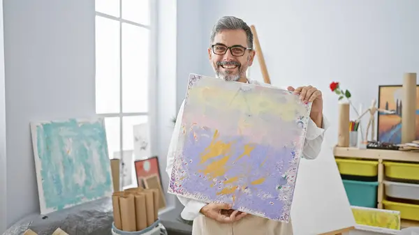 Joyful young hispanic male artist with grey-haired beard, wearing glasses and apron, confidently showing his splendid painting at the art studio, smiling beamingly with pure enjoyment and creativity
