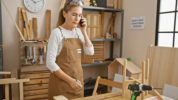 Blonde woman talks on phone multitasking in a sunny carpentry workshop with wood and tools.