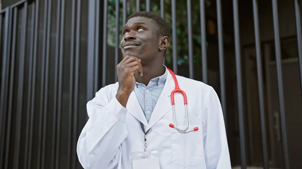A thoughtful african man in a white lab coat with a stethoscope standing outside a hospital, looking up.