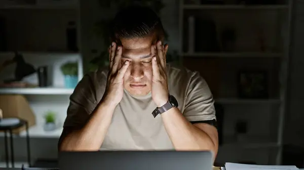 Stressed latin man tired of overwork at his desk, seriously tackling business hurdles with laptop in dim office