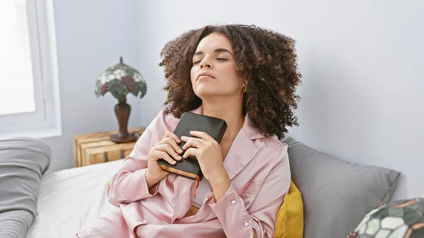 A young, attractive hispanic woman with curly hair dressed in pink, holding a wallet while sitting in a modern bedroom interior.
