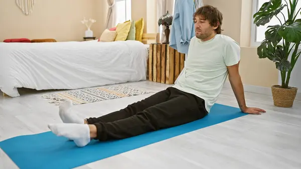 A young man seated on a yoga mat in a cozy bedroom, looking relaxed and contemplative