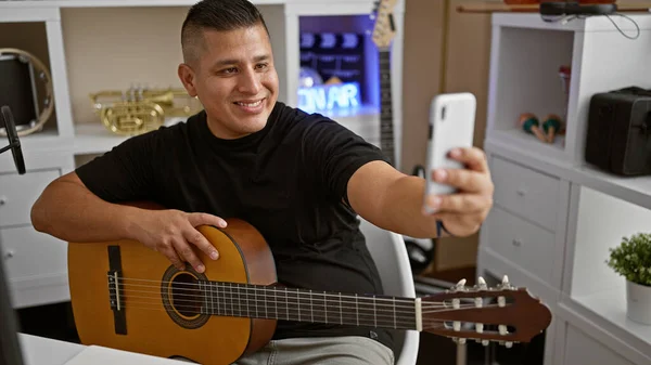 笑着的年轻拉丁裔男子 一个年富力强的艺术家 在他的古典吉他上谱曲 在一个音乐工作室里拿着智能手机自拍 — 图库照片