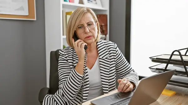 Зрелая Женщина Офисе Разговаривает Телефону Работает Ноутбуке Изображая Напряженную Профессиональную — стоковое фото