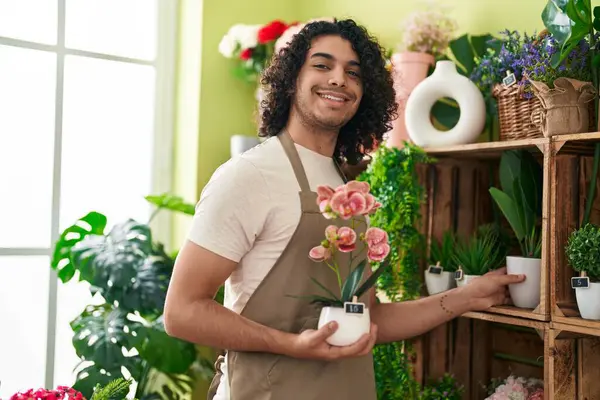 Young latin man florist smiling confident holding plant at flower shop