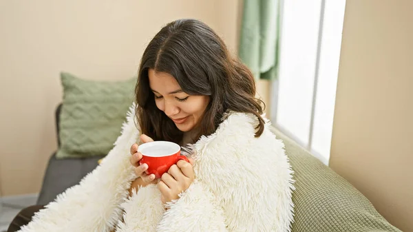 A serene hispanic woman enjoys a warm beverage at home, exuding a cozy and relaxed atmosphere in her living room.