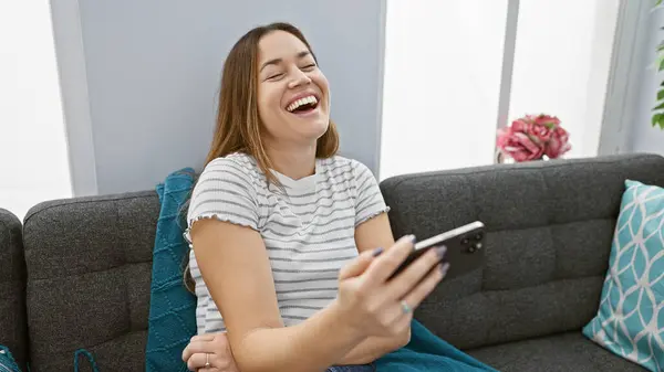 Laughing woman holding smartphone on grey sofa in modern living room.