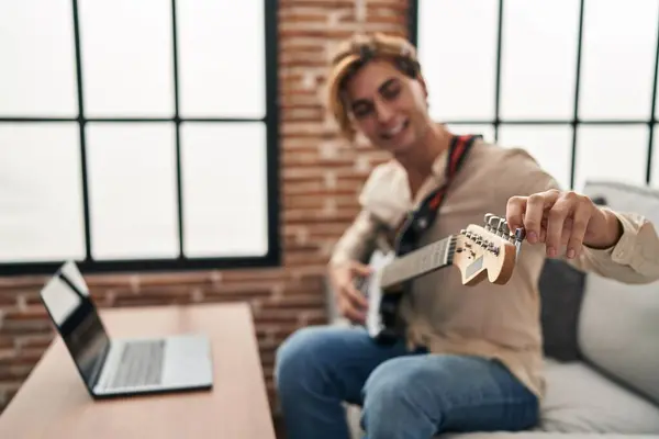 Young caucasian man having online electrical guitar class sitting on sofa at home