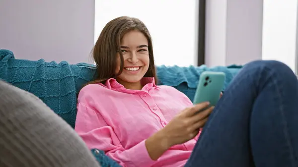 楽しい若いヒスパニック女性 幸せを放射し リビングルームのソファに座って 電話で陽気なメッセージを入力し 自宅でオンラインインタラクションを楽しむ — ストック写真