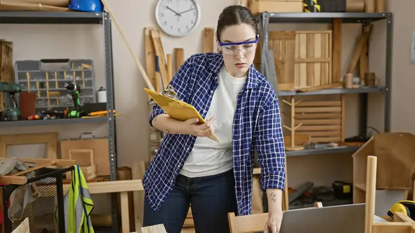 stock image Caucasian woman inspects clipboard in a workshop filled with woodworking tools and furniture.