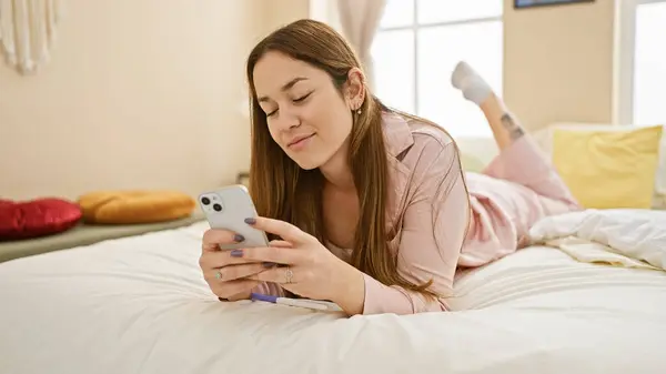 年轻女人躺在舒适的卧室里 用智能手机展示着随意的家庭生活方式 — 图库照片
