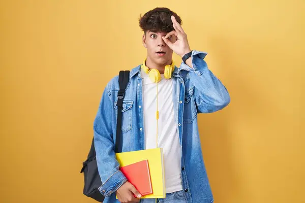 Hispanic teenager wearing student backpack and holding books doing ok gesture shocked with surprised face, eye looking through fingers. unbelieving expression.