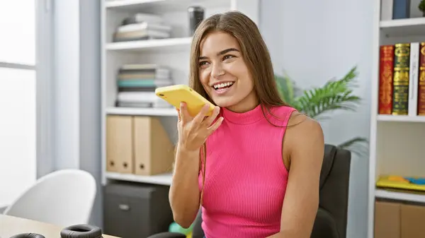 Empowered young hispanic business woman confidently sends voice message from her smartphone, radiating success while working in her sleek office
