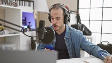 Müzik stüdyosunda sakallı, kulaklık takan ve akıllı telefon kullanan Latin kökenli kel bir adam..