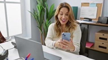 Modern ofiste akıllı telefon kullanan gülümseyen İspanyol kadın, iş arkadaşlarıyla bir video görüşmesi sırasında ekranda mutlu bir şekilde el sallıyor..
