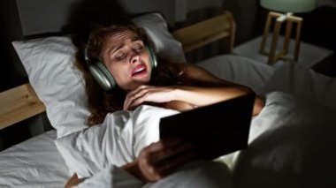 Kulaklıklı, genç, beyaz bir kadın yatakta duygusal bir şekilde uzanıp tableti evde izliyor..