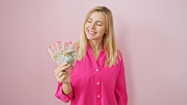 喜ばしい若い女性 ブロンドと自信を持って 孤立したピンクの背景の上に偶然に新しいゼーランドドルを保持しています 成功のポジティブで歯止めの笑顔 — ストック動画