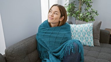 Düşüncelere dalmış genç bir kadın, gri bir kanepede, rahat bir oturma odasında battaniyeye sarılmış olarak oturuyor..