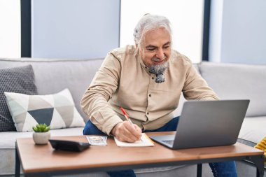 Evde dizüstü bilgisayar kullanan orta yaşlı, kır saçlı bir adam.