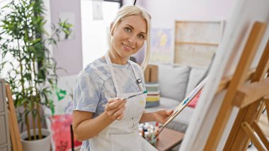 Mutlu genç sarışın kadın sanatçı, kendine güvenen ve gülümseyen, üniversite sanat stüdyosunda resim dersinin keyfini çıkarırken, boya fırçalarıyla güzel tablolar çiziyor.
