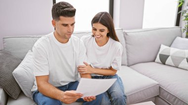 Gülümseyen bir kadın ve odaklanmış bir adam modern bir oturma odasında bir kanepeye oturup belgeyi birlikte inceliyorlar..