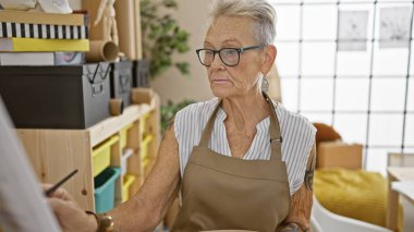 Olgun, gri saçlı kadın sanatçı sanatına tamamen dalmış, özenle kendi stüdyosunda çizim yapıyor, yaratıcılığı ve konsantrasyonu somutlaştıran sessiz bir insan.