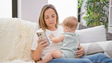 Rahat anne, kanepede oturuyor, akıllı telefonuyla mesajlaşırken ciddi bir ifade takınıyor, küçük kızıyla evde güzel bir anı paylaşıyor.