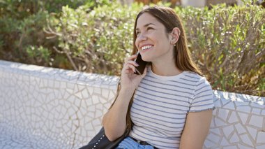 Bir telefon görüşmesinde gülümseyen genç bir kadın, güneşli bir parkta oturmuş, yeşillik içinde, günlük rahatlama ve bağlantıyı somutlaştırıyor..