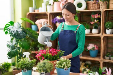 Young beautiful hispanic woman florist smiling confident watering plant at flower shop