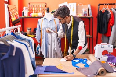 Young hispanic man tailor writing on notebook holding shirt at atelier
