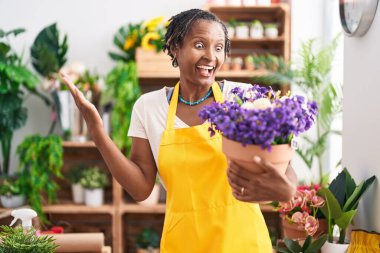 Çiçekçi dükkanında çalışan Afrikalı bir kadın elinde çiçekle başarıyı kutluyor mutlu bir gülümseme ve elini kaldırarak kazanan bir ifade. 