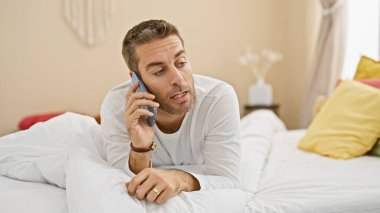 Yakışıklı sakallı adam rahat bir yatak odası ortamında telefonda uzanıyor, evde rahatlık ve iletişim tasvir ediyor..