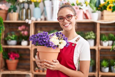 Çiçekçi dükkanında çalışan genç İspanyol bir kadın elinde bitkiyle mutlu ve pozitif görünüyor. Dişlerini gösteren bir gülümsemeyle ayakta duruyor. 