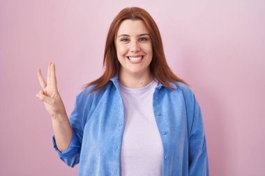 Pembe arka planda duran ve üç numaralı parmaklarıyla işaret eden, kendine güvenen ve mutlu gülümseyen İspanyol genç kadın.. 