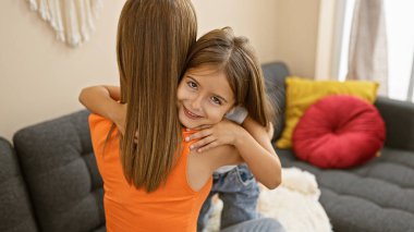 Anne ve kızı sıcak bir oturma odasında sevgi dolu bir şekilde kucaklaşıyorlar, aileyi, şefkati ve sade bir atmosferi resmediyorlar..