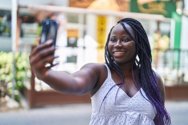 Afro-Amerikalı kadın gülümsüyor. Sokakta akıllı telefonun yanında özçekim yapıyor.