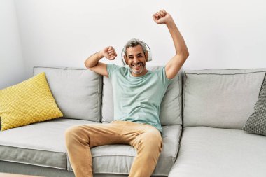 Orta yaşlı, kır saçlı, dans eden ve müzik dinleyen bir adam. Evdeki kanepede oturuyor.