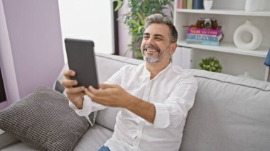 Gri saçlı, neşeli İspanyol bir adam gülümsüyor ve dokunmatik yastıkta eğlenceli bir video görüşmesi yapıyor. Oturma odasındaki kanepesinde dinleniyor.