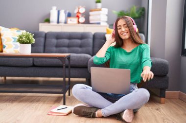 Young beautiful hispanic woman sitting on floor listening to music and dancing at home