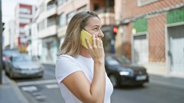 Şık genç sarışın kadın ciddi telefon konuşmalarına dalmış, güneşli şehir caddesinde dikiliyor, konsantrasyonunu gösteriyor, gelişigüzel, kentsel bir yaşam tarzını kusursuz bir şekilde yakalıyor..