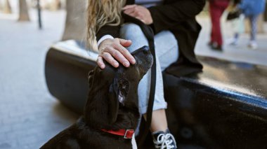 Sarışın kadın şehir caddesinde siyah labradoru okşar, şehir yaşamının ortasında huzurlu bir anı yakalar..