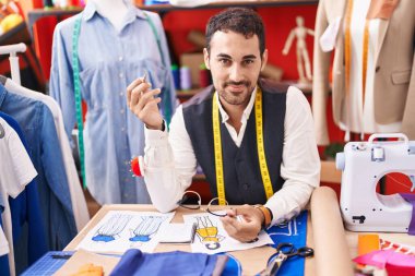 Young hispanic man tailor smiling confident sitting on table at atelier
