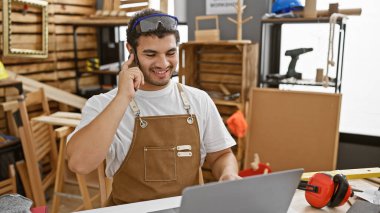 Güvenlik gözlüğü takan, sakallı ve atölye önlüklü gülümseyen bir adam marangoz stüdyosunda telefonla konuşuyor..
