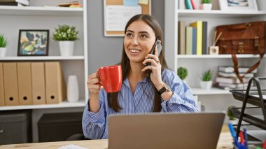 Gülümseyen genç bir İspanyol kadın modern bir ofiste elinde kırmızı bardakla telefon görüşmesi yapıyor..