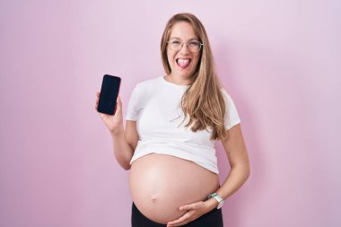Genç hamile bir kadın karnını gösteriyor. Akıllı telefonu tutuyor. Dilini dışarı çıkarıyor.. 