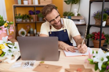 Middle age man florist using laptop writing on notebook at flower shop