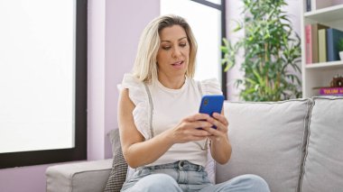 Kanepede oturan akıllı telefon kullanan genç sarışın kadın ciddi bir yüzle evde oturuyor.