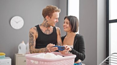 Çamaşır odasında çamaşır sepetine yaslanmış akıllı telefon kullanan güzel bir çift.