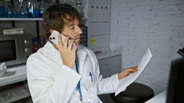 Laboratuvar önlüklü beyaz bir adam elinde bir belge tutarken akıllı telefonla konuşuyor..
