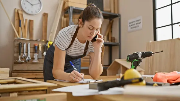 Focused Woman Carpentry Workshop Talks Phone While Taking Notes Amidst — Stock Photo, Image